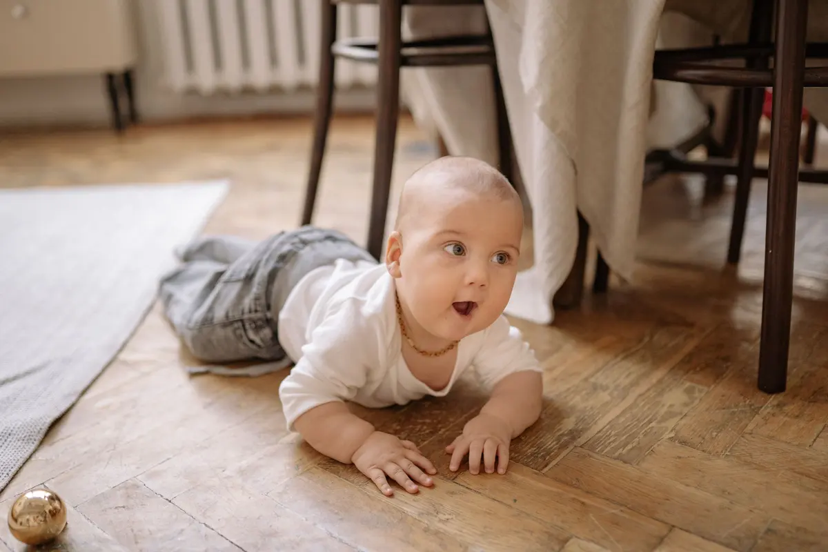A baby in a white shirt crawling across a herringbone wood floor beside a dining table